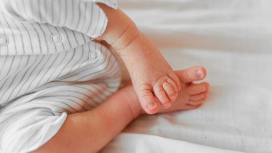 Cute sleeping newborn baby feet on white blanket. Baby in a white gender neutral romper on the bed. Little toes, heels.