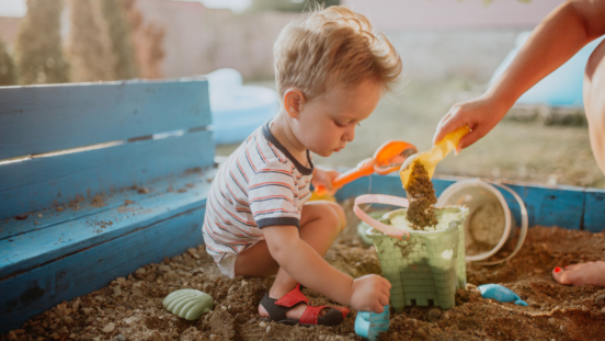 Blonde haired todler boy crouching and playing in a sand pit