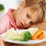 Fair haired boy with his head on one arm with plate of broccoli, carrots and potatoes in front of him