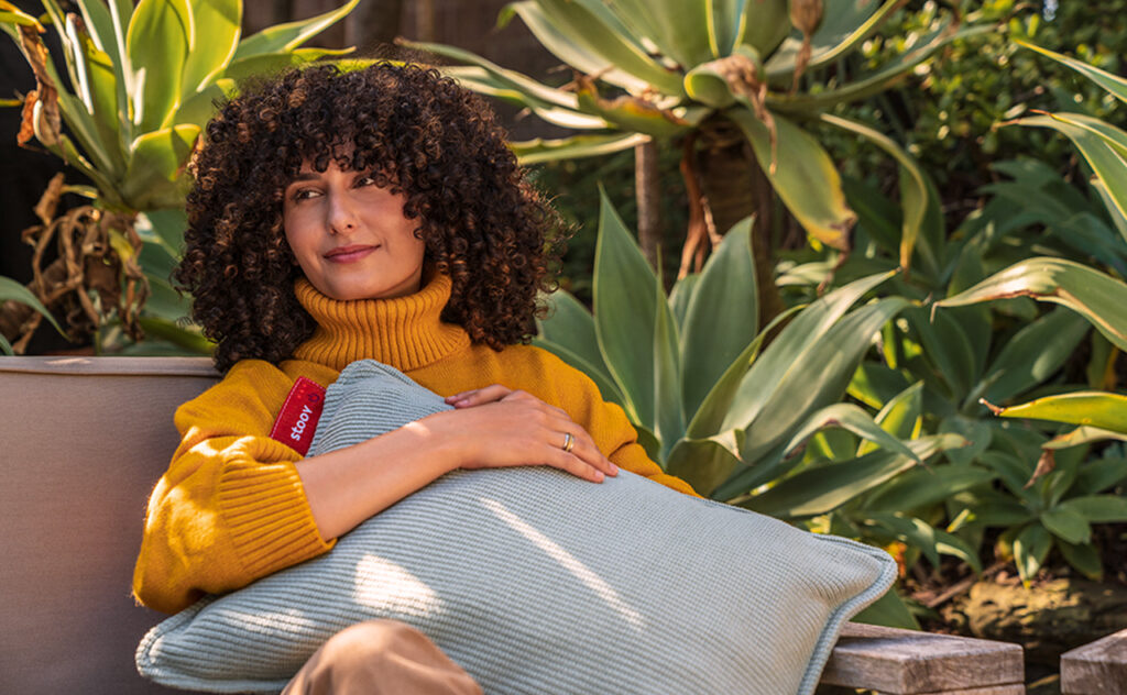 STOOV Ploov Cordless Heating Cushion lifestyle image of curly haired woman hugging cushion to her chest while sitting outdoors