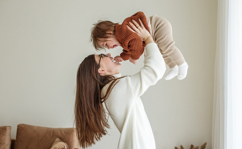 Happy cheerful young mother throws an adorable baby in air, plays, hugs baby with love, care, enjoys motherhood, maternity leave. Family spends time together.