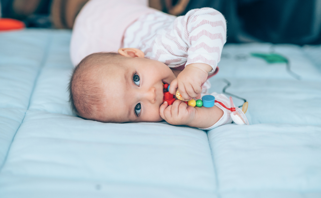 Cute baby dressed in pink lying on her side with a wooden teether toy in her mouth, looking towards the camera