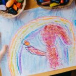 Child's crayon drawing of a rainbow in a blue sky and a pony tailed child with an orange butterful on their hand