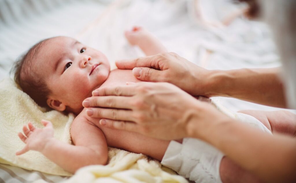 Relaxed Asian baby wearing a nappy lies with their arms up by their head as a pair of hands massages them