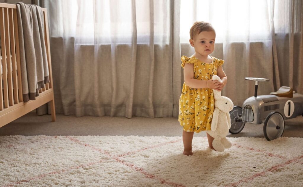 Toddler girl in cute yellow sleeveless all-in-our carries a toy bunny by its ears, walking barefoot on a lovely carpet with sheet curtains in the background