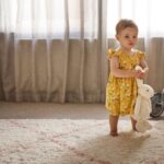 Toddler girl in cute yellow sleeveless all-in-our carries a toy bunny by its ears, walking barefoot on a lovely carpet with sheet curtains in the background