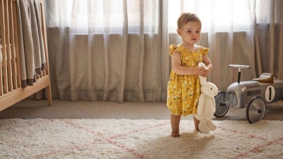 Toddler girl in cute yellow sleeveless all-in-our carries a toy bunny by its ears, walking barefoot on a lovely carpet with sheet curtains in the background