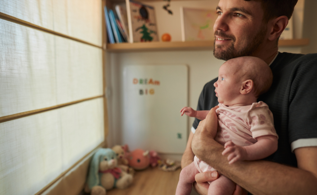 Man with dark beard holding a young baby as they look towards the window