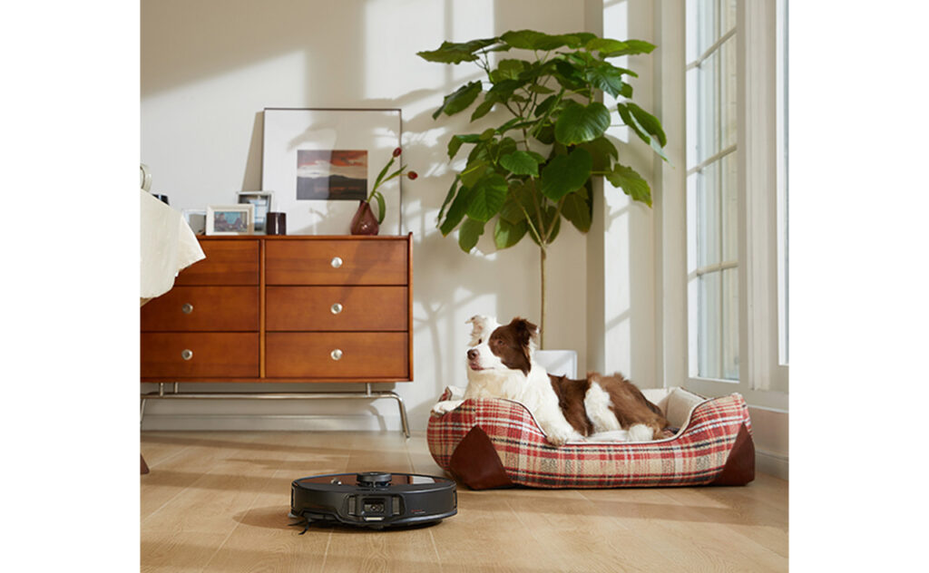 Roborock S8 MaxV Ultra lifestyle shot of vac working in room while a reclining border collie looks on.