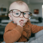 Blonde baby wearing a brown jumper, fingers in mouth, wearing adult glasses