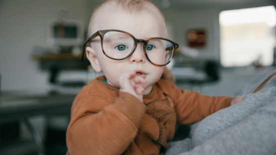 Blonde baby wearing a brown jumper, fingers in mouth, wearing adult glasses