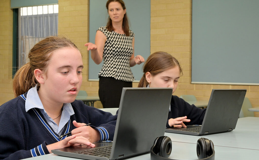 Technology teacher(Female age 30-40) teaching schoolgirls how to use laptops in school classroom.
