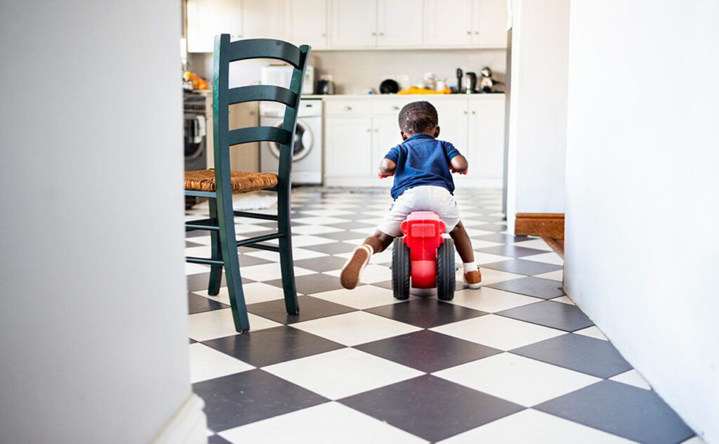 Rear view of a cute little African boy riding his toy tricycle around his kitchen at home