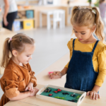 Toddler girl with ponytail in a Montessori style classroom looking at t green tray with an older girl with blonde bunches