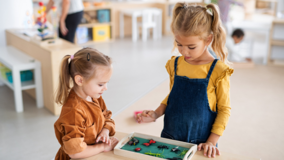 Toddler girl with ponytail in a Montessori style classroom looking at t green tray with an older girl with blonde bunches