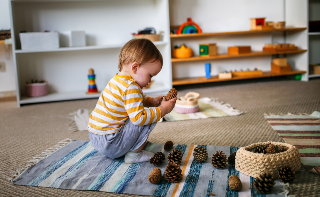 Toddler in yellow striped top playing on the floor surrounded by pine cones