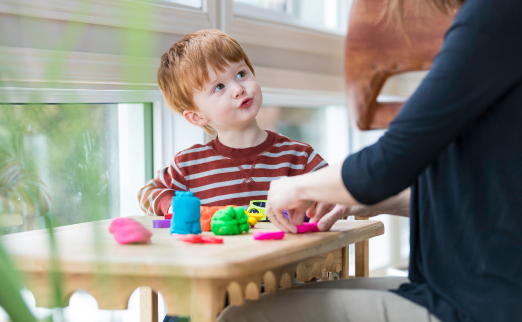 Red-head five year old boy in front of colourful playdoh looking at an adult in a Montessori style classroom