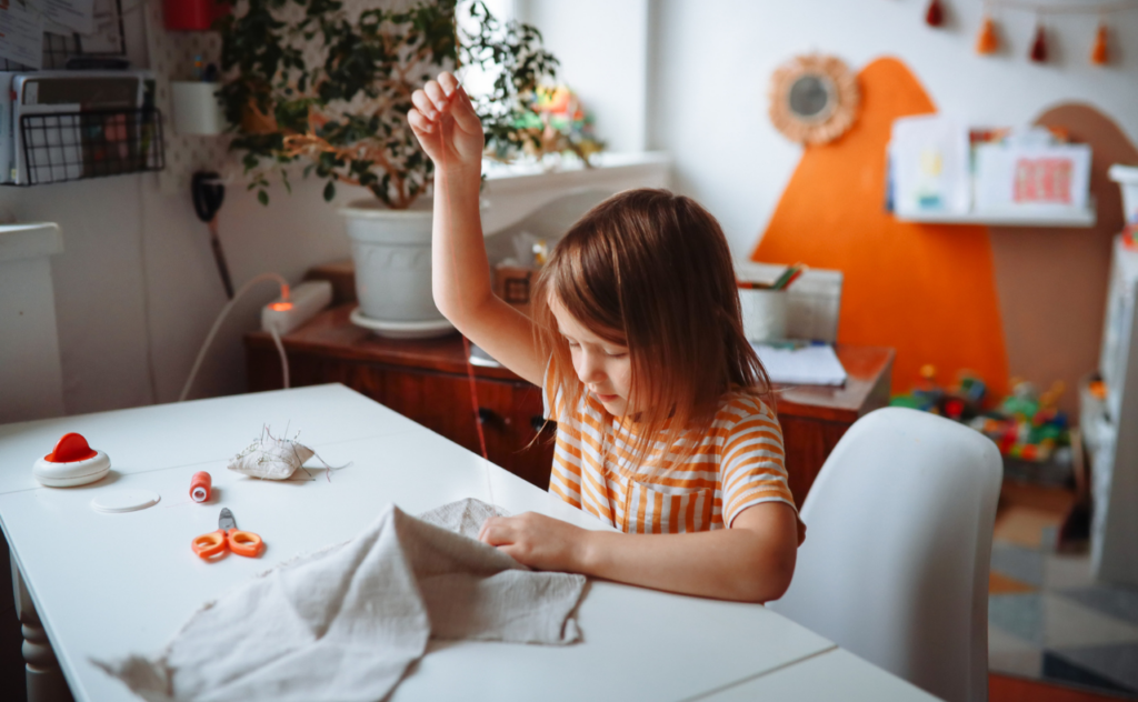 Primary school aged girl in a striped orange top, hand high in the air as she sews a piece of fabric in a Montessori class