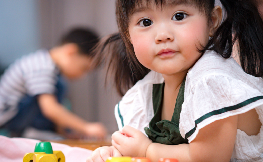 Cute Asian toddler girl with bangs and bunches looks at camera, coloured Montessori building blocks