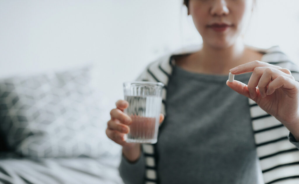 Young Asian woman taking medicines with a glass of water at home, with a pill bottle by the side.