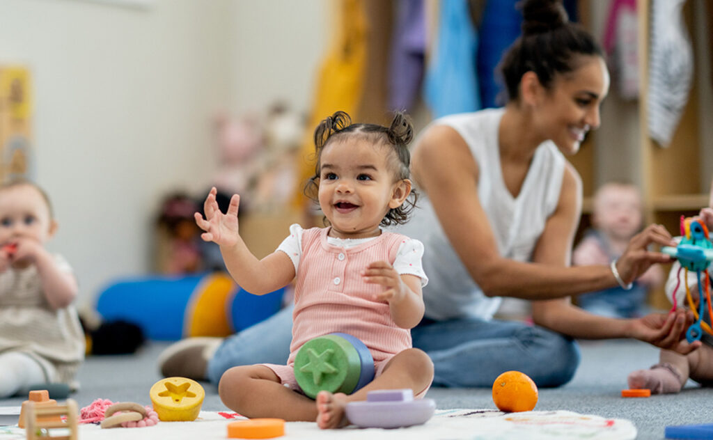 A small group of babies sit on the floor playing with various toys while their teacher supervises and engages them. They are each dressed casually and appear content.