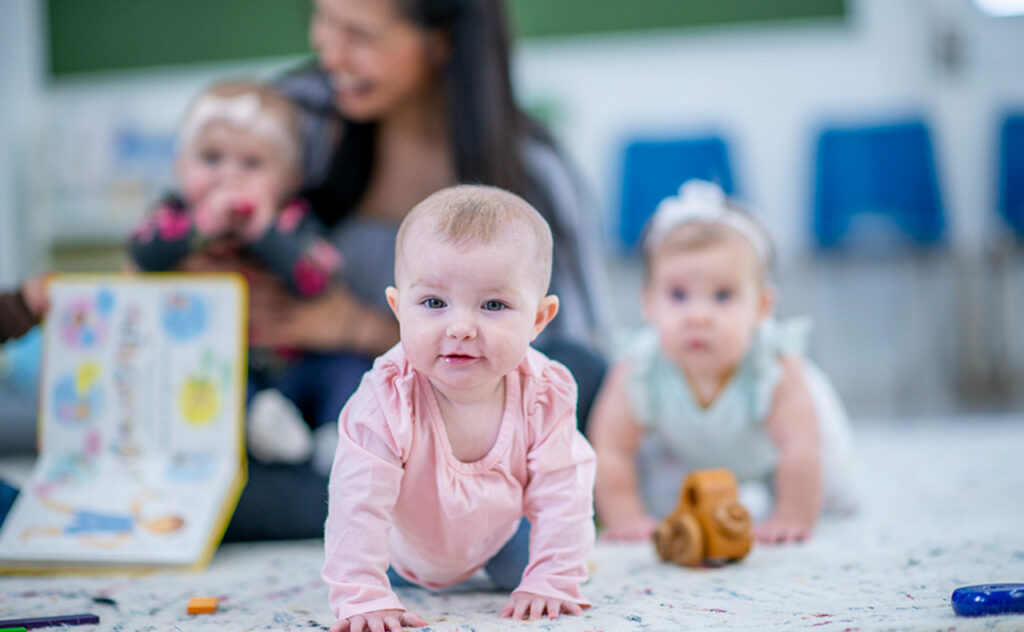 A little Caucasian girl crawls among other multi-ethnic babies on a daycare room floor. She is smiling and enjoying her pay time. A daycare worker is sitting in the background with another little girl on her lap. There are toys and storybooks out around the room.