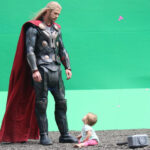 Chris Hemsworth in thro costume against green wall while baby daughter looks up at him