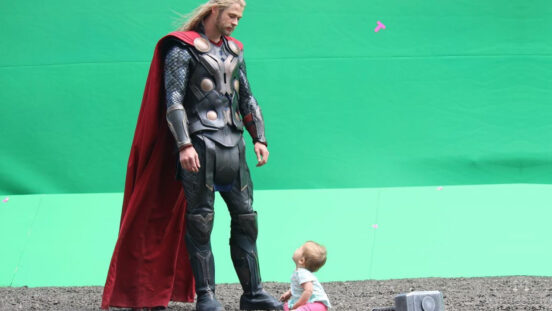 Chris Hemsworth in thro costume against green wall while baby daughter looks up at him