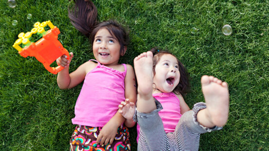 Two toddler girls lie on grass laughing as they play with bubble machine.