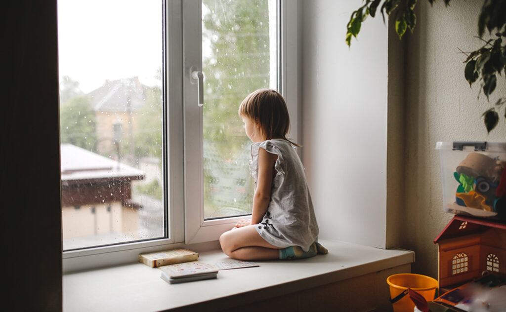 One toddler girl with short haircut sits on windowsill and looks out window with rain.