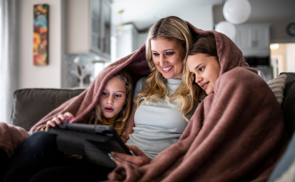 Blonde hair woman sitting on a sofa with two kids cuddled up with a pale pink blanket around their shoulders