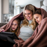 Blonde hair woman sitting on a sofa with two kids cuddled up with a pale pink blanket around their shoulders