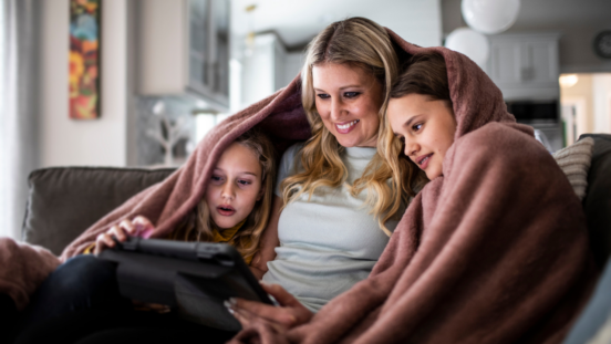 Blonde hair woman sitting on a sofa with two kids cuddled up with a pale pink blanket around their shoulders