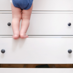 Chubby bare legs of toddler using open drawers like steps to climb up a piece of furniture