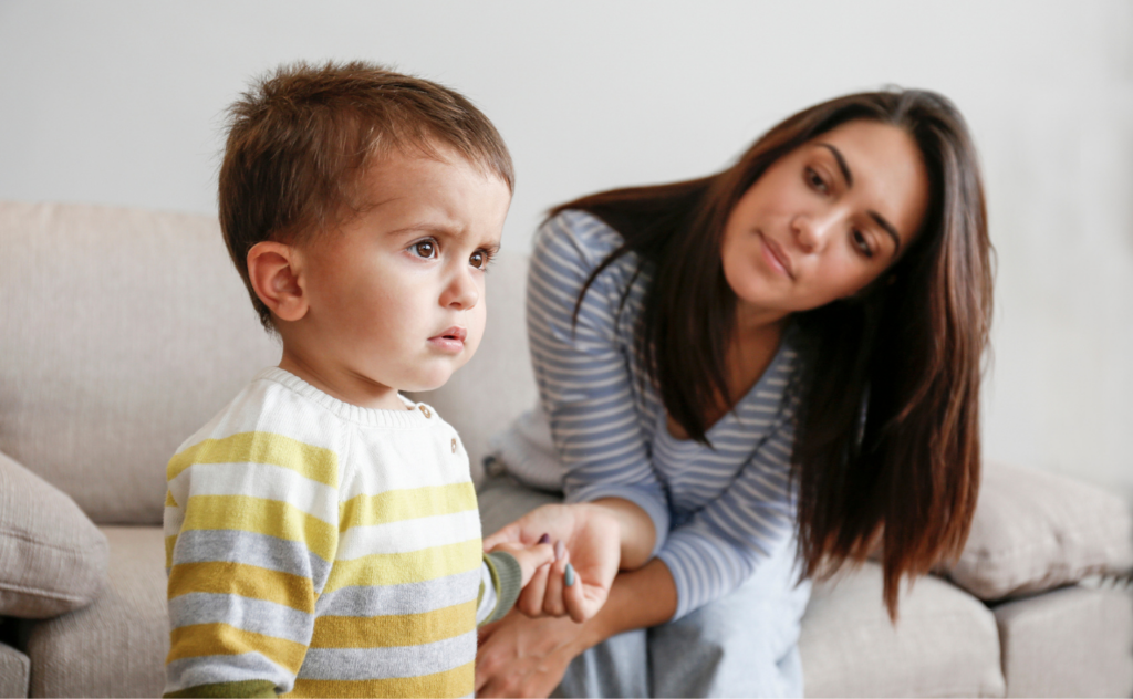 Mum with long dark hair sitting on a sofa holding her toddler son's hand as he looks away from her, not listening