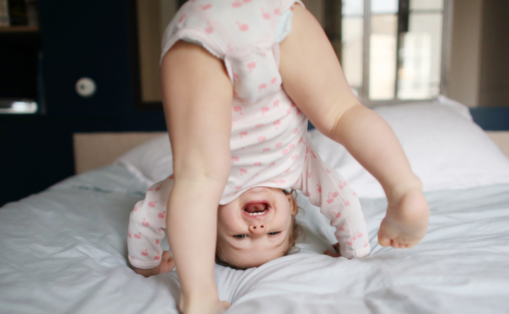 Older baby wearing pink and standing her head laughing as she looks through her legs towards the camera