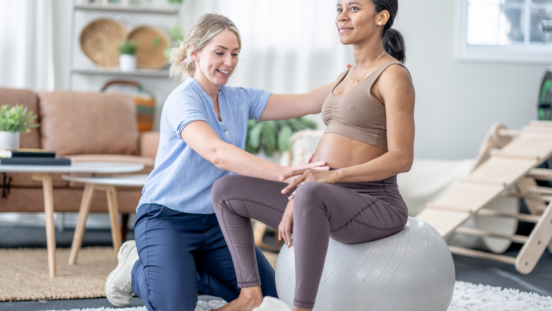 Pregnant woman wearing sports gear, sitting on a yoga ball while a woman in casual scrubs kneels next to her with her hand on her belly