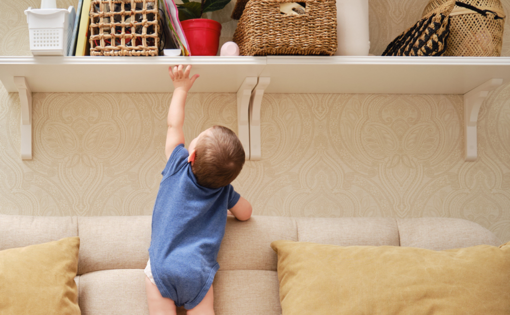 Young toddler in blue t-shirt climbing on the sofa to reach up to the high shelf