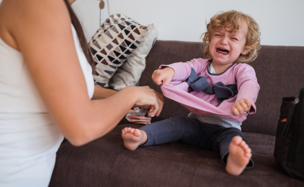 Toddler girl sitting on a sofa, screaming and pulling at her top as her mum tries to put socks on her