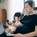 Pregnant Asian woman looking at her laptop while a toddler girl looks at a tablet in the background