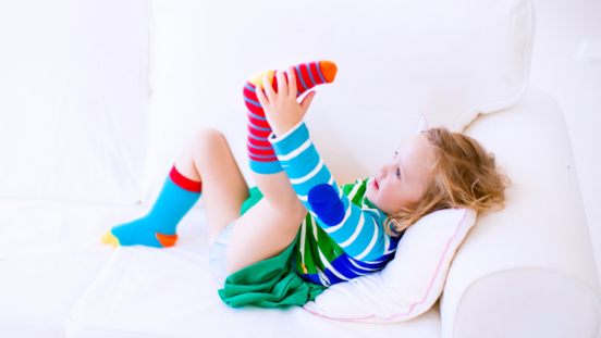 Fair-haired toddler girl wearing brightly coloured clothes lying on her back on a pale sofe with her feet in the air