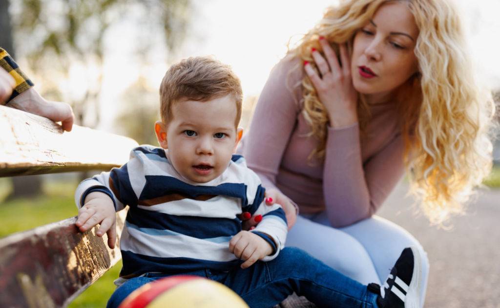 Mum sitting on park bench holding a toddler's arm as he looks away not listening to her