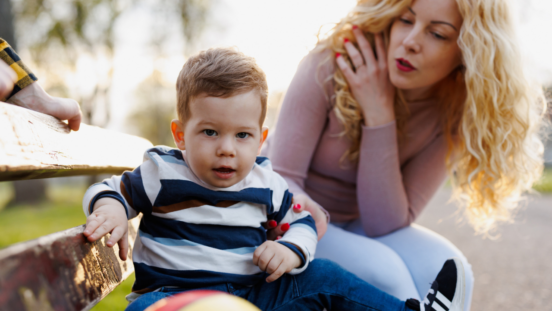 Mum sitting on park bench holding a toddler's arm as he looks away not listening to her