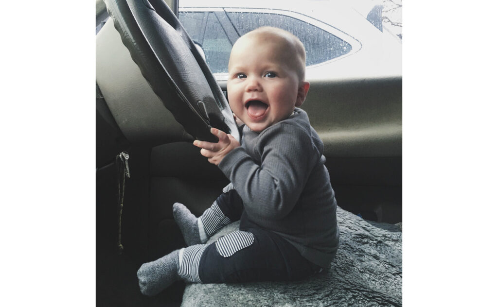 Happy young baby sitting on the drivers seat of car holding onto the steering wheel
