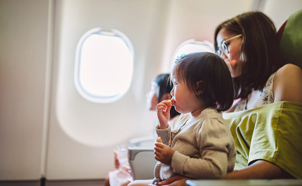 Little toddler girl having snacks joyfully while sitting on her mum’s lap on the airplane