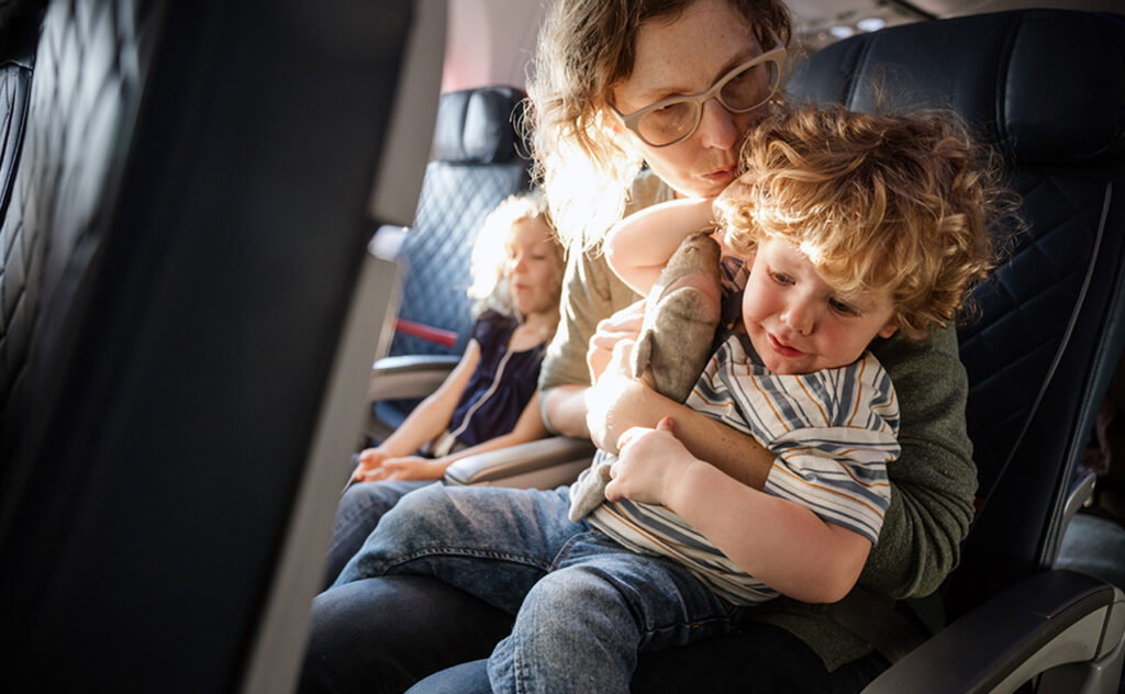 A Caucasian family with multiple children on vacation depart from the airport on a plane. The mother holds a crying child experiencing ear pain on the flight.