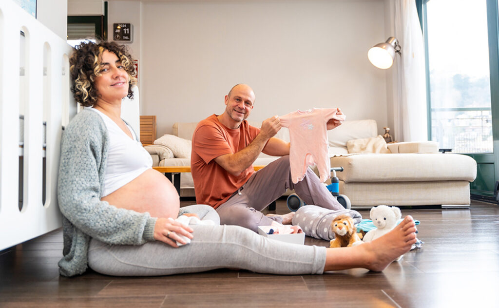 Happy adult and caucasian pregnant parents preparing baby clothes sitting on the ground at home
