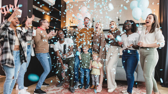 Diverse group of adult close friends cheering and smiling as the blue confetti reveal the gender of the baby at the gender reveal party. The expectant couple is having a baby boy. They all look happy. The confetti are flying everywhere and the apartment is a mess. Fun and heartfelt moment.