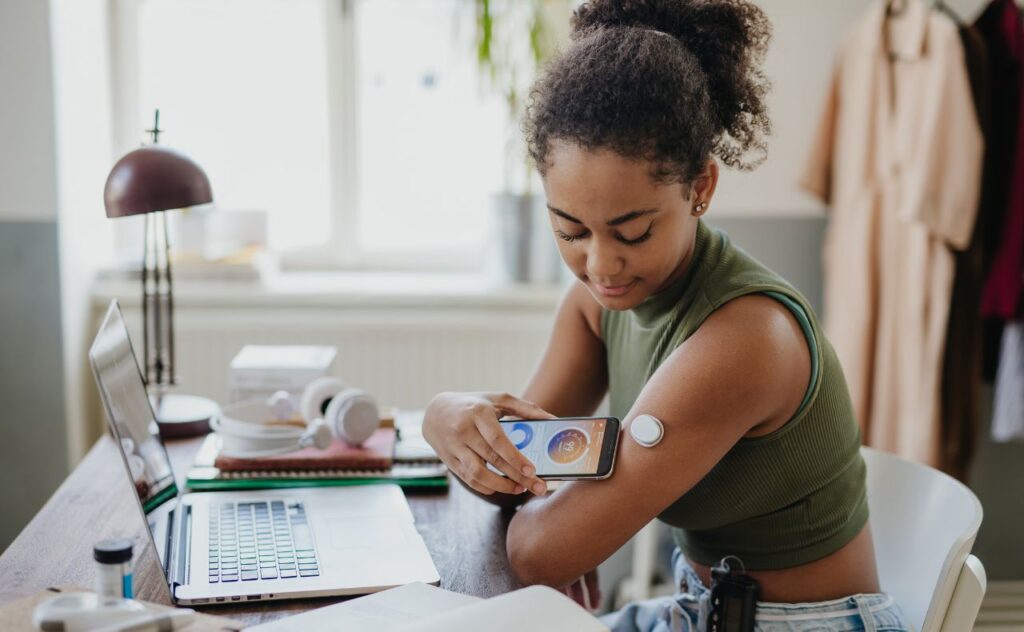 Teenage girl connecting smartphone to continuous glucose monitor, checking blood glucose during day. 