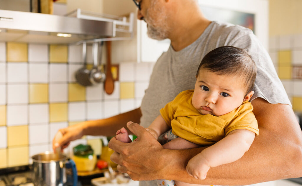 Middle aged man cooking in a kitchen while holding a three months old baby on the other hand